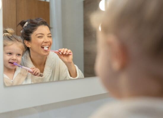 Mother and toddler girl in bathrobes brushing their teeth together in front of a bathroom mirror