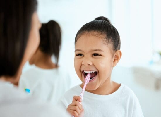 Mother helping her young daughter brush teeth correctly while both look into the bathroom mirror
