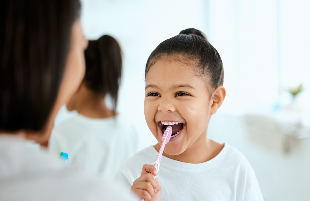 Mother helping her young daughter brush teeth correctly while both look into the bathroom mirror